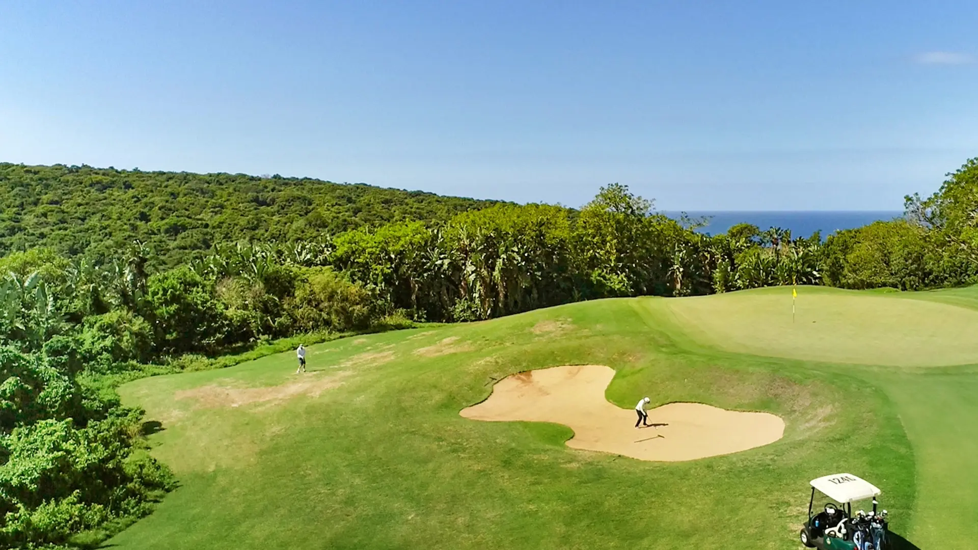 Aerial view of a fairway with a bunker in the middle. Trees are off to the left and the blue ocean is in the distance